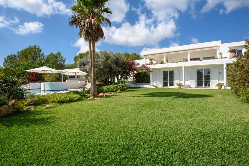 A luxurious white villa, perfect for luxury travel, boasts multiple balconies set in a lush green garden under a blue sky. A swimming pool surrounded by sun umbrellas and palm trees enhances the serene and inviting atmosphere. David Pearson Travel