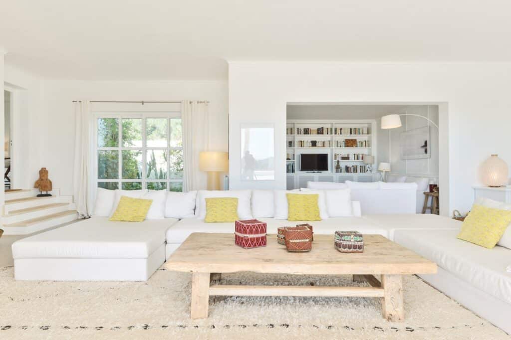 A spacious, bright living room exudes a touch of luxury travel with its white walls and furniture. A large wooden coffee table adorned with decorative items sits at the center. Yellow pillows accent the white couches, while large windows and a built-in bookshelf complete this chic retreat. David Pearson Travel