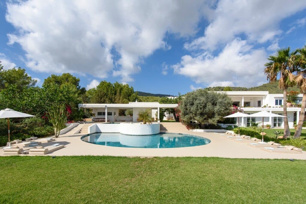 Experience luxury travel at its finest in this spacious modern villa, featuring a large swimming pool surrounded by sun loungers and umbrellas. Lush greenery, palm trees, and scenic hills provide a breathtaking backdrop under a blue sky dotted with scattered clouds. David Pearson Travel