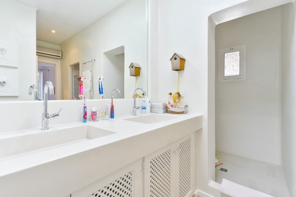 The bright, modern bathroom offers a touch of luxury travel with double sinks and a large mirror. The counter holds various toiletries, while a small, windowed shower is visible to the right. Crisp white walls and vanity create a clean, airy atmosphere reminiscent of upscale retreats. David Pearson Travel