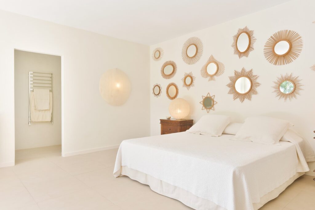 A minimalist bedroom exuding luxury travel vibes features a large bed draped in white linens. The wall boasts an array of sunburst mirrors, while a wooden dresser stands to the right. Through an open doorway on the left, a towel rack is visible, and soft, warm lighting enhances the serene ambiance. David Pearson Travel