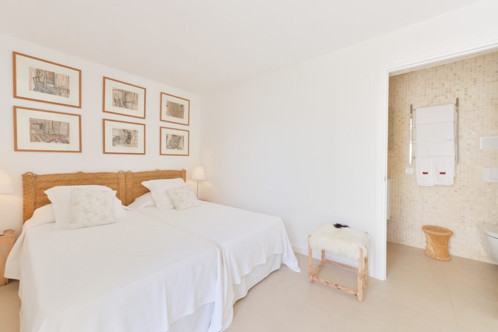 A bright bedroom epitomizing luxury travel, featuring two single beds with white linens and a wicker headboard. A small wooden table sits between them, and six framed pictures adorn the wall above. An open door reveals a bathroom with tiled walls and plush towels on a rack. David Pearson Travel