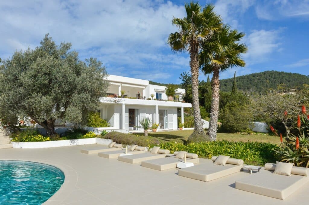 A modern white villa epitomizes luxury travel, featuring a spacious terrace with cushioned lounge chairs beside a curved swimming pool. Lush greenery, palm trees, and a large olive tree surround the area, with hills visible in the background under a partly cloudy sky. David Pearson Travel