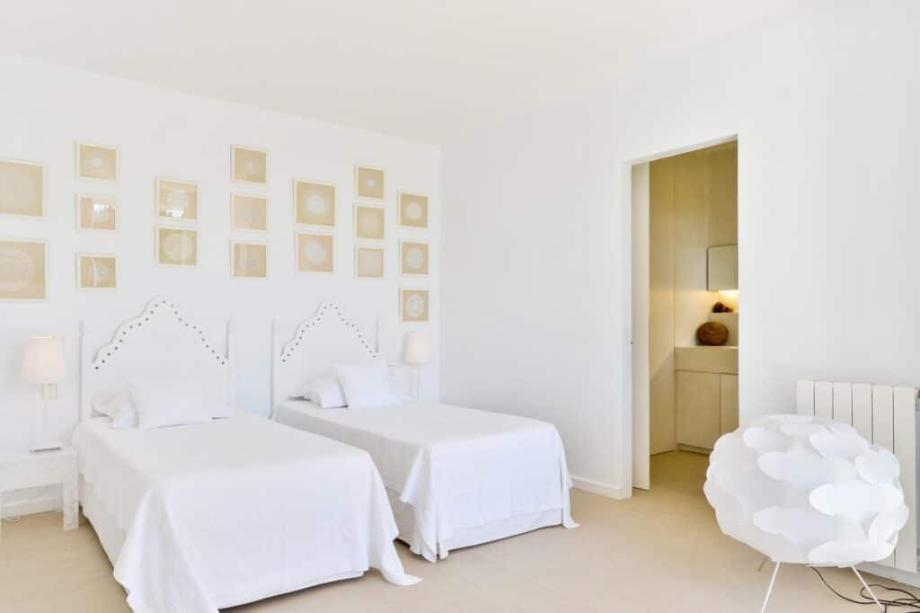 A minimalist bedroom with two twin beds, each adorned with white linens and unique headboards, offers a touch of luxury travel. Above is a grid of framed art. Soft lighting creates a serene atmosphere. A modern white chair sits nearby, while an open door reveals a glimpse of the bathroom. David Pearson Travel