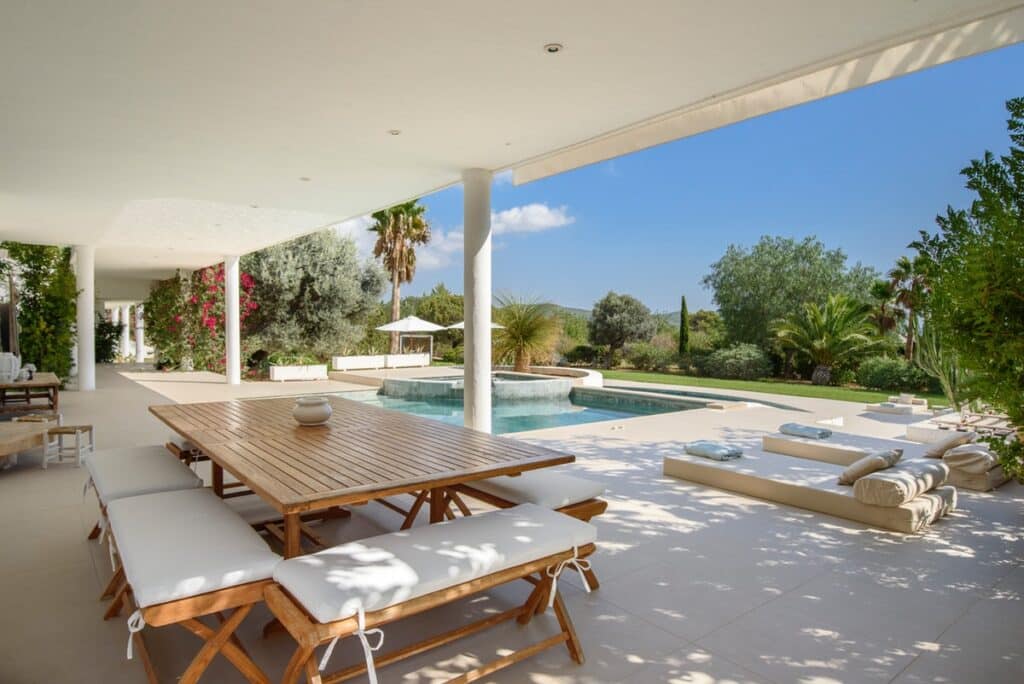 Spacious outdoor patio with a wooden dining table and cushioned benches, perfect for luxury travel experiences. Overlooks a swimming pool surrounded by lush greenery, including palm trees and flowering plants, all under a clear blue sky. David Pearson Travel