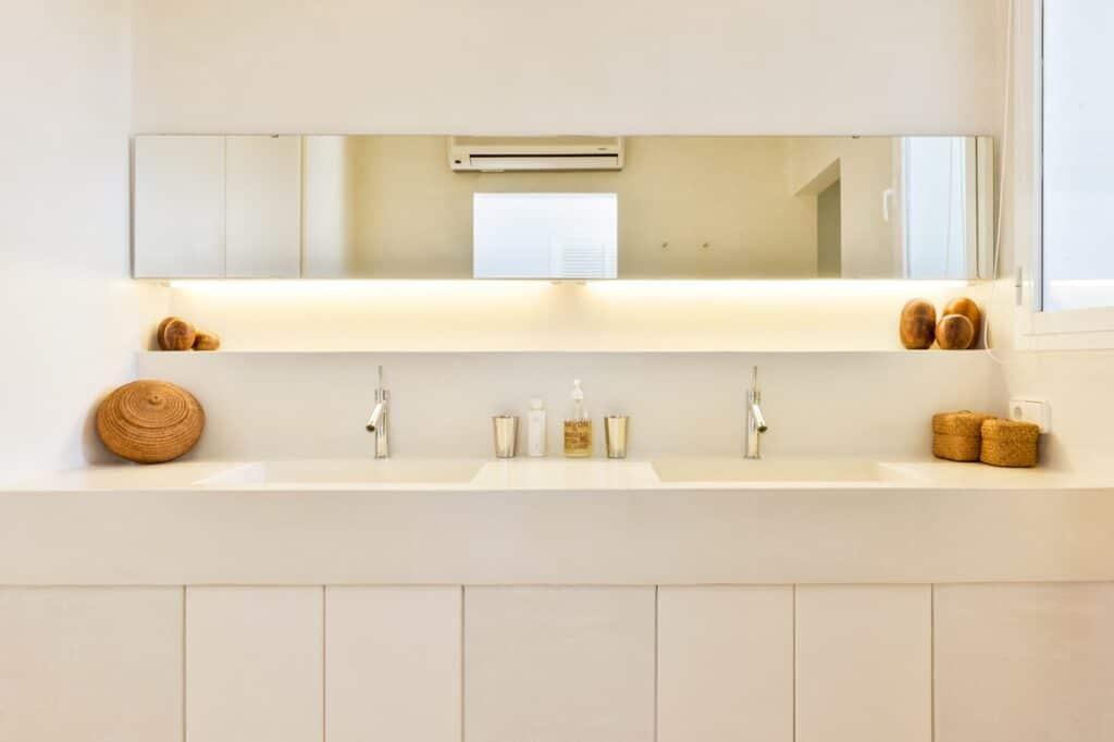 A modern bathroom evokes luxury travel with its dual sinks on a sleek white countertop. Above, a long mirror with warm, backlit lighting enhances the ambiance. Decorative wicker baskets and jars sit neatly below the wall-mounted air conditioner, completing this elegant retreat. David Pearson Travel