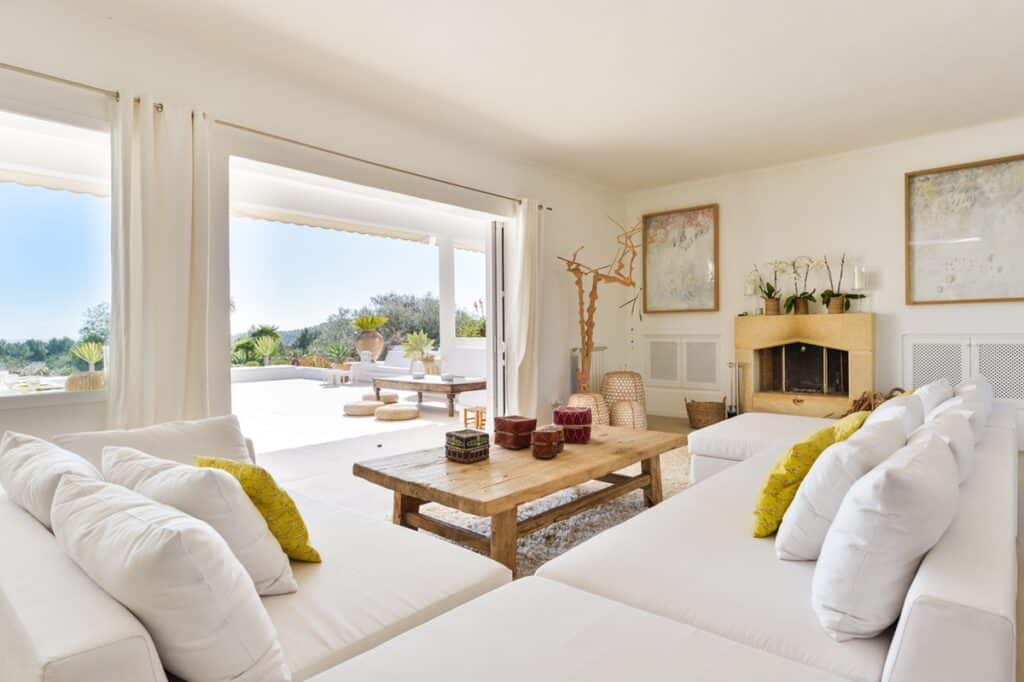 A bright luxury living room with large windows opening to a patio. It features white sofas, yellow pillows, and a wooden coffee table. A fireplace is on the right, and the room flourishes with plants and abstract art. Natural light fills this oasis, reminiscent of a luxury travel retreat. David Pearson Travel