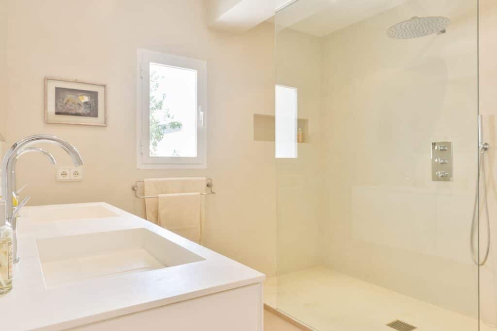 A bright, modern bathroom exuding luxury travel vibes features a white double sink vanity with sleek faucets, a large glass-enclosed shower, and a window. A towel is neatly hung on a rack, and a small framed picture adds charm to the wall. David Pearson Travel