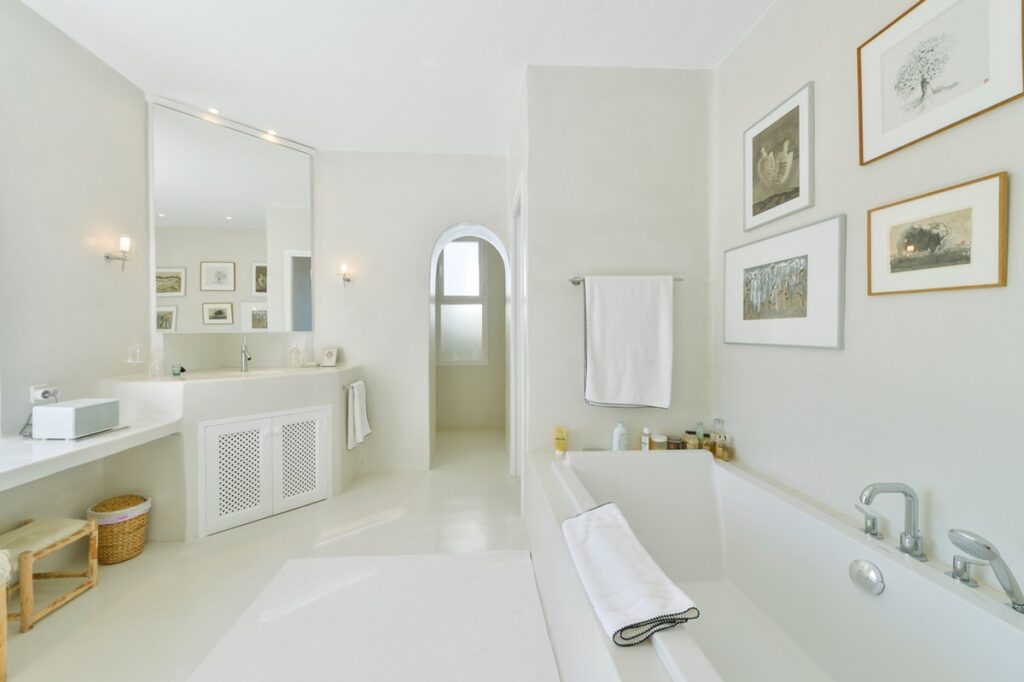 Experience a touch of luxury travel in this spacious, minimalist bathroom featuring a large white bathtub, pristine white walls, and light floor tiles. Elegant decor includes framed art, a mirror above a sink with a plush white towel, and a wicker basket. Soft lighting enhances the serene atmosphere. David Pearson Travel