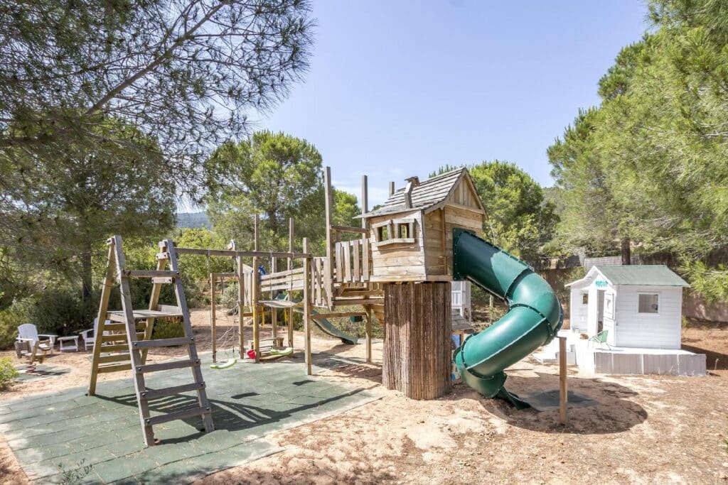 A playground surrounded by trees features a wooden play structure with a green spiral slide, swings, and a small white playhouse. The ground is covered with lush sand and green rubber tiles reminiscent of a luxury travel retreat. The scene is set under a clear blue sky. David Pearson Travel
