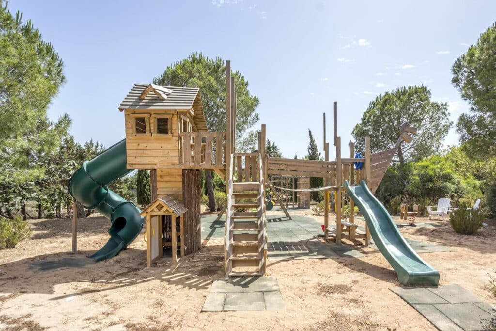 A luxury travel destination for kids, this wooden playground set in a park features a treehouse, rope bridge, and two slides—one green and spiral. Encircled by trees, it rests on sandy surfaces with rubber mats under a clear blue sky. David Pearson Travel