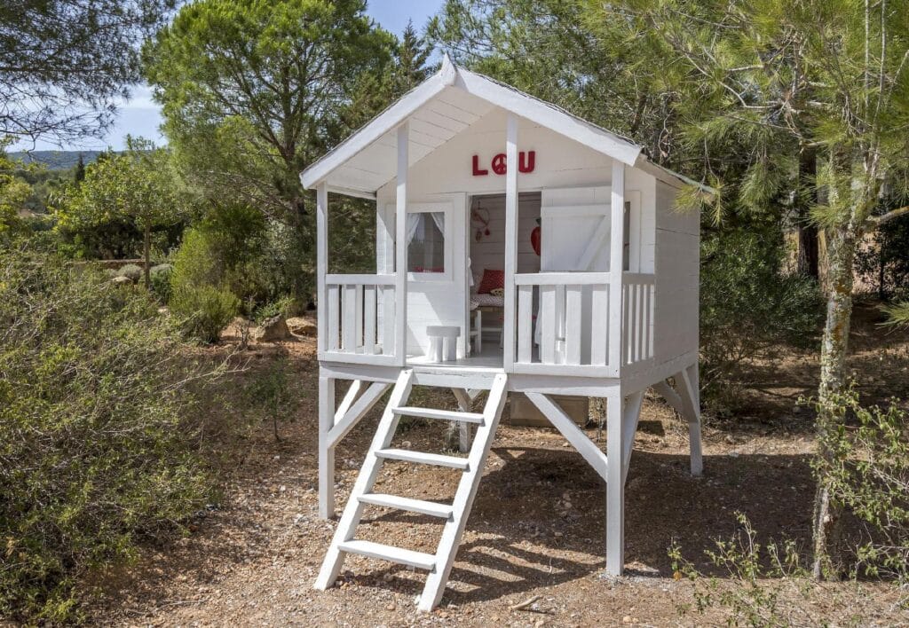 Nestled in a forested area, a charming white wooden playhouse on stilts invites explorers. A ladder leads to its entrance, windows overlook the greenery, and a quaint porch adds to its allure. Above the door, LOU is inscribed in red letters—nature’s luxury travel experience for the imagination. David Pearson Travel