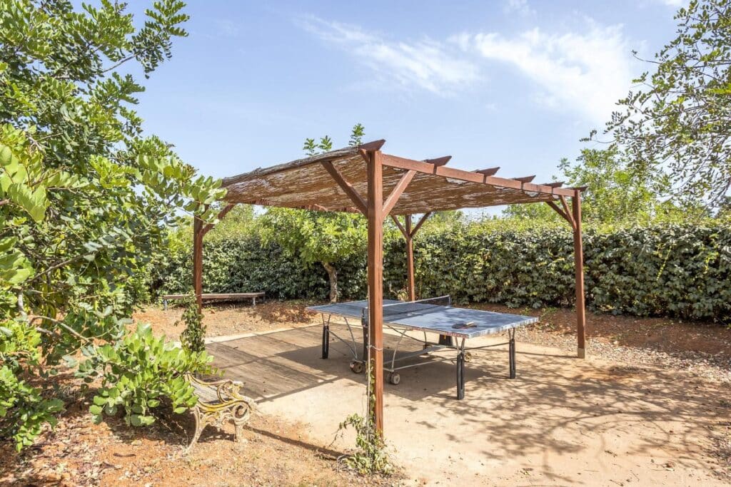 A ping pong table sits elegantly under a wooden pergola in a sunny outdoor setting, perfect for those savoring luxury travel. The area is surrounded by lush greenery and a dirt ground, while a wooden bench provides relaxation nearby under the clear sky dotted with a few clouds. David Pearson Travel