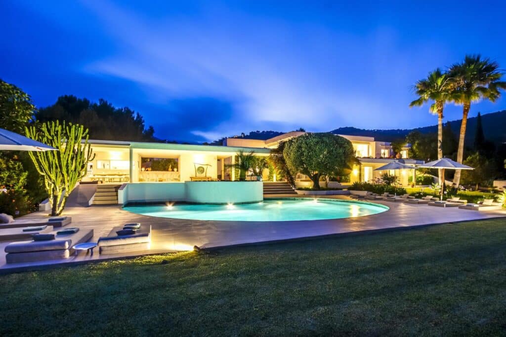 Luxurious modern house with a curved pool in the foreground, perfect for a luxury travel destination, surrounded by deck chairs and umbrellas. Lush greenery and palm trees in the background, all set against a twilight sky. David Pearson Travel