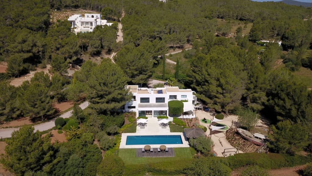 Aerial view of a white modern villa, epitomizing luxury travel, surrounded by lush greenery and trees. The villa features a rectangular pool with sun loungers and outdoor seating. A winding path leads to another similar villa in the background, promising an exquisite escape. David Pearson Travel