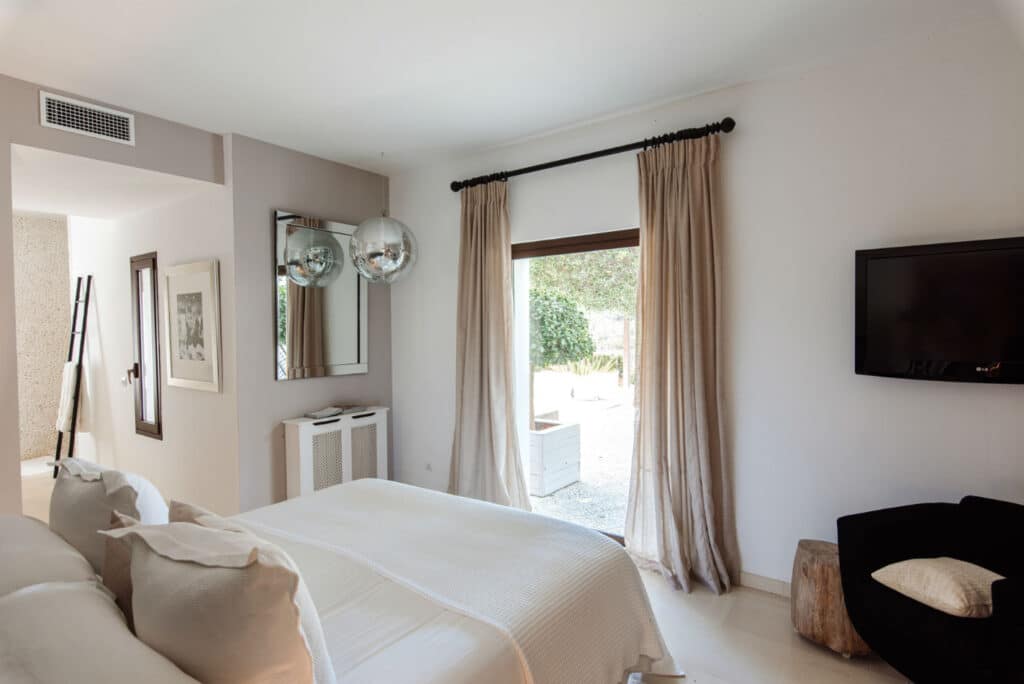 A modern bedroom exuding the elegance of luxury travel, featuring a large bed with beige bedding and a wall-mounted TV. Two silver hanging lamps and a mirror adorn a small console. The spacious window reveals stunning outdoor views, complemented by a black chair and decorative ladder. David Pearson Travel