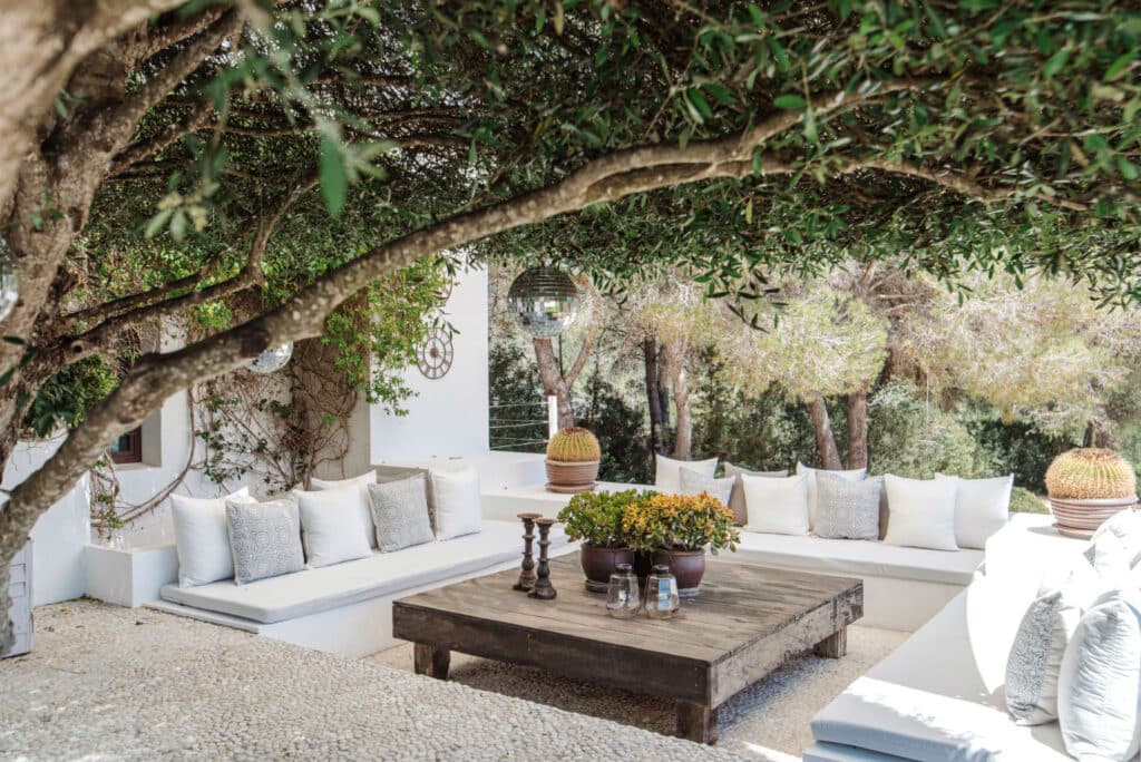 Experience luxury travel right in your backyard with a cozy outdoor seating area featuring white cushions, a large wooden coffee table, and lush decorative plants under a canopy of dense trees. Surrounded by serenity and vast greenery on a textured stone floor, its pure tranquility. David Pearson Travel