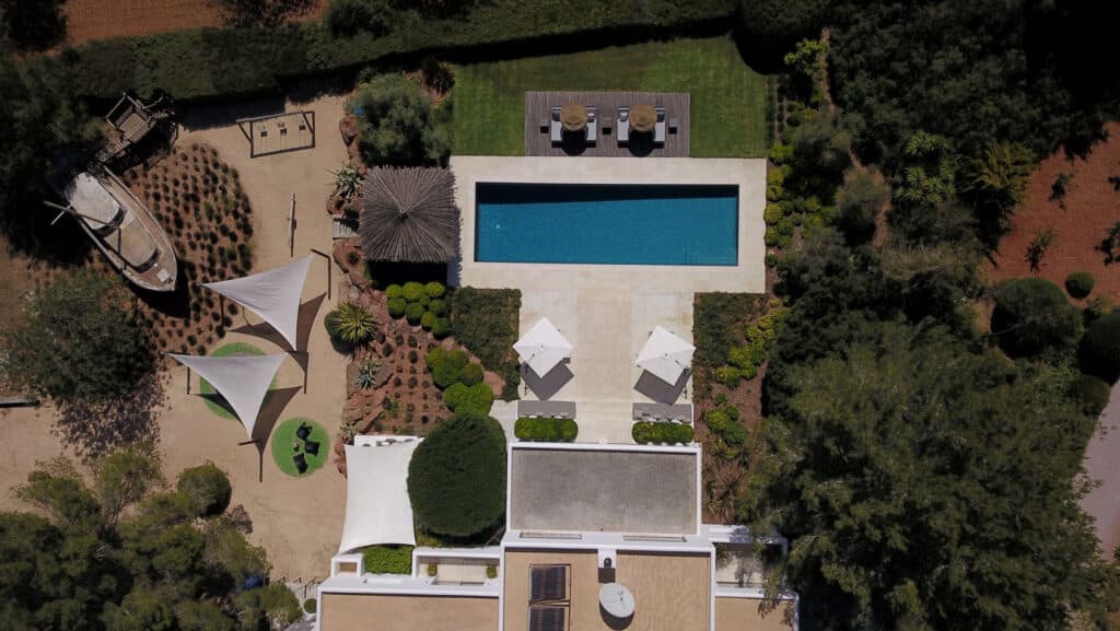 Aerial view of a landscaped backyard featuring a rectangular pool surrounded by lush greenery. This luxury travel oasis includes umbrellas and seating, a boat-shaped structure, a playground, tents, and a small round table with chairs set on sandy ground for an exquisite outdoor experience. David Pearson Travel