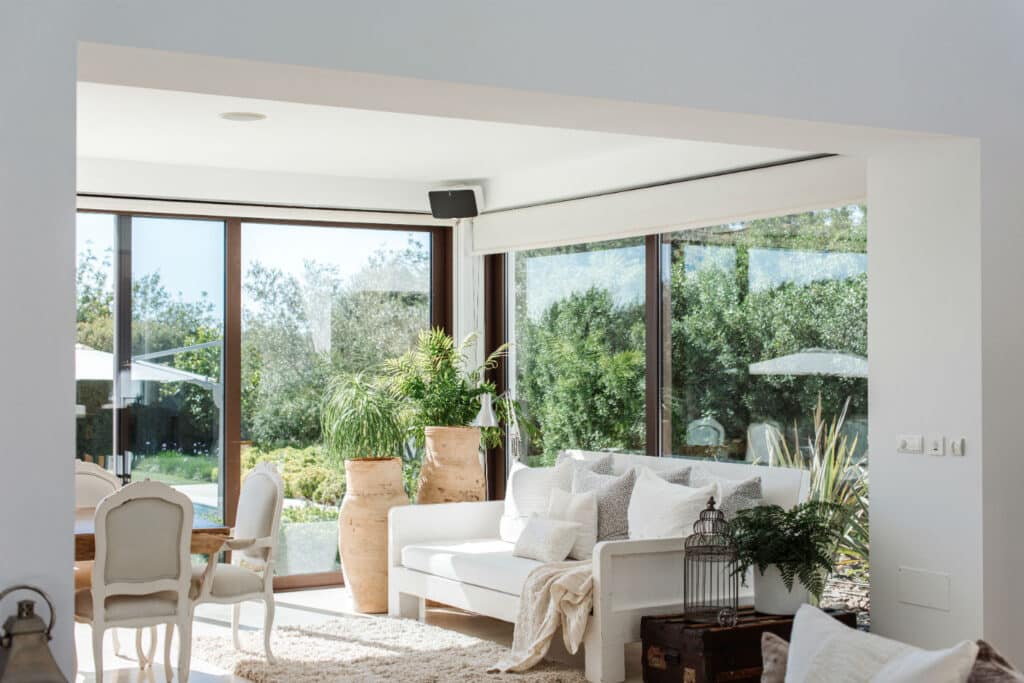 Bright living room with large windows showcasing greenery outside, reminiscent of a luxury travel retreat. Inside, a white sofa with cushions, potted plants, a birdcage, and a wooden dining table with chairs create an airy and light decor that exudes a cozy, inviting atmosphere. David Pearson Travel