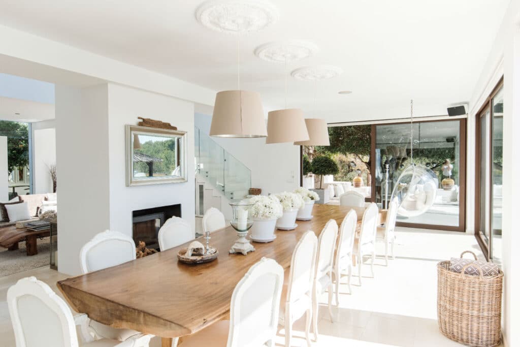 Bright dining room with a long wooden table and white upholstered chairs. Large windows offer garden views reminiscent of luxury travel settings. Three pendant lights hang above the table, and a round swing chair is visible in the background. Décor includes white flowers and a mirror. David Pearson Travel