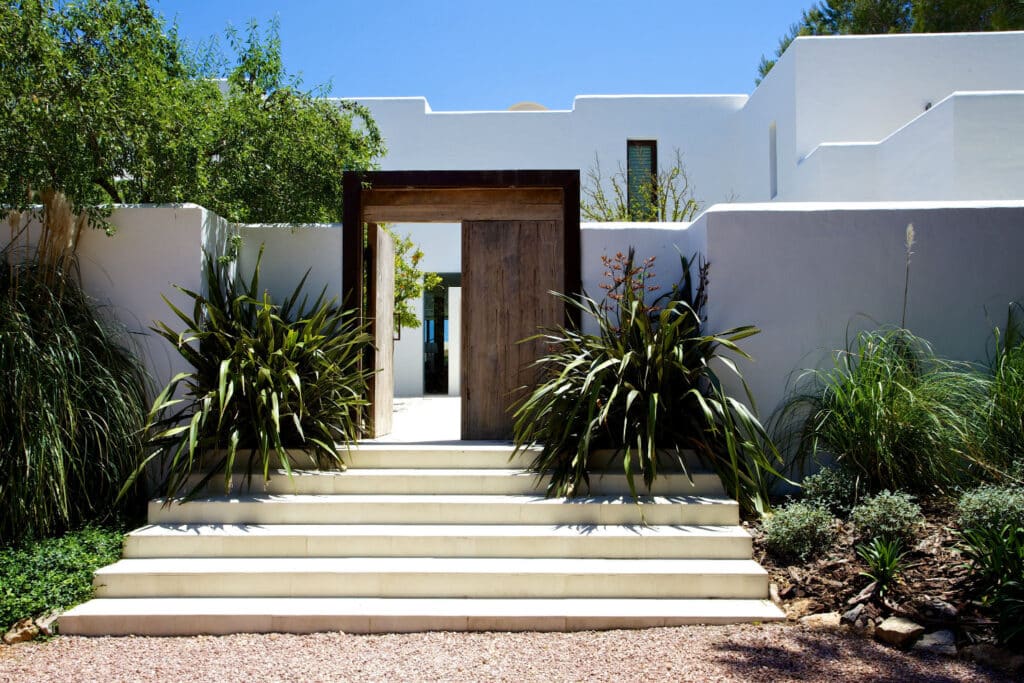 The luxury travel-inspired exterior of this modern Mediterranean-style house boasts pristine white stucco walls. A wooden door frame and set of stairs welcome you, flanked by lush green plants, all set against a backdrop of clear blue sky. David Pearson Travel