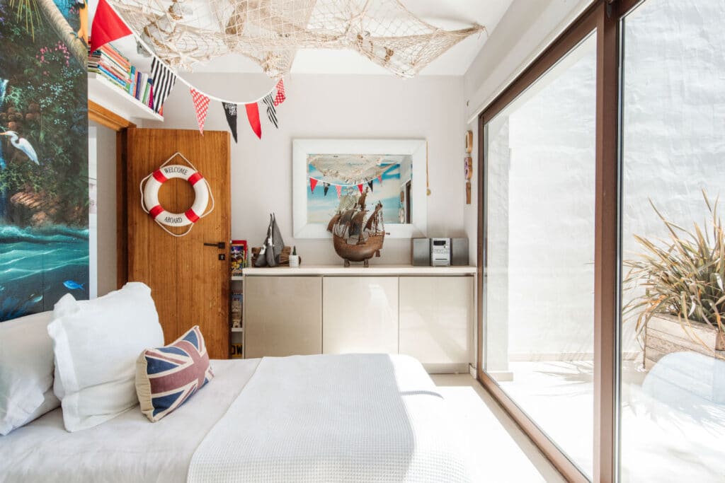 A brightly lit bedroom exuding luxury travel vibes with its nautical decor. It features a bed adorned with a Union Jack pillow and a lifebuoy on the wooden door. A model ship, decorative fishing net, and vibrant red and white bunting complete the scene through the large window. David Pearson Travel