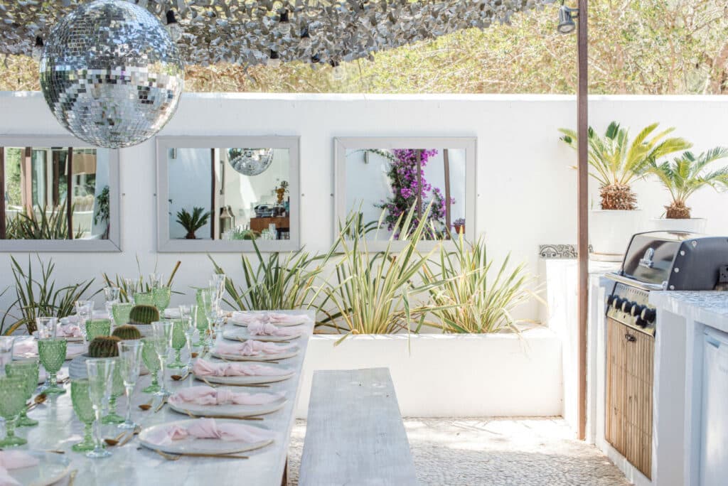 The luxurious outdoor dining area features white decor and a patterned stone floor. A long, elegant table is adorned with pink napkins and green glassware. Above, a large disco ball glistens; mirrors and plants embellish the walls, while a sleek grill completes this upscale travel experience. David Pearson Travel