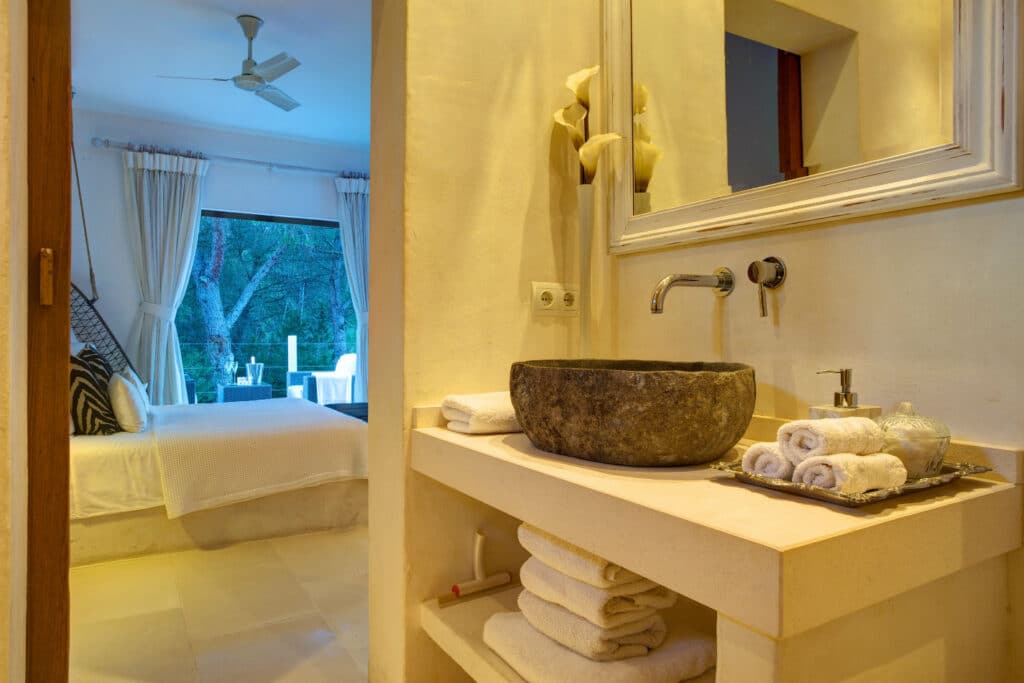 A cozy bathroom with beige walls and a stone sink on a counter, adorned with rolled towels, exudes the charm of luxury travel. In the background, discover a bedroom featuring white bedding, curtains, and a scenic view through the window. A ceiling fan hangs above. David Pearson Travel