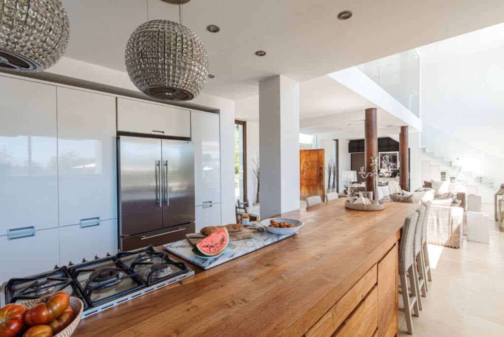 The modern kitchen exudes the charm of luxury travel with its wooden island, sleek stainless steel fridge, and a gas stovetop. Spherical pendant lights cast a soft glow on a slice of watermelon and croissants, while stairs lead to an elegantly designed living area in the background. David Pearson Travel