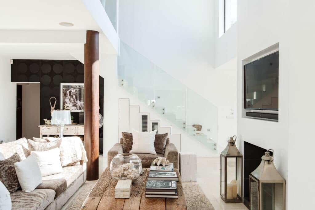 A modern, sunlit living room exuding luxury travel vibes with a large beige sofa, wooden coffee table, and a mounted TV above the fireplace. A glass staircase in the background and decorative items evoke elegance, while tall candles in lanterns on the floor enhance the cozy ambiance. David Pearson Travel