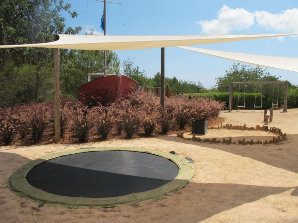 A playground featuring a sunken trampoline amidst luxurious landscaping, surrounded by sand. A red boat rests in the background with lush plants nearby, complementing the sophisticated swing set. Elegant white sunshades provide shelter, while distant trees and bushes complete this travel-worthy retreat. David Pearson Travel