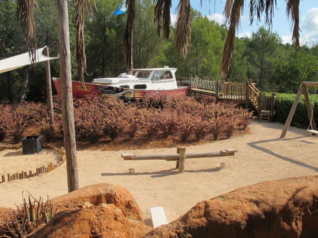 The playground, perfect for a luxury travel retreat, features a seesaw and swings over sandy terrain. A red and white boat sits elegantly amidst ornamental grasses, with palm leaves providing shade and trees as a scenic backdrop. A charming bridge invites exploration of the boat area. David Pearson Travel