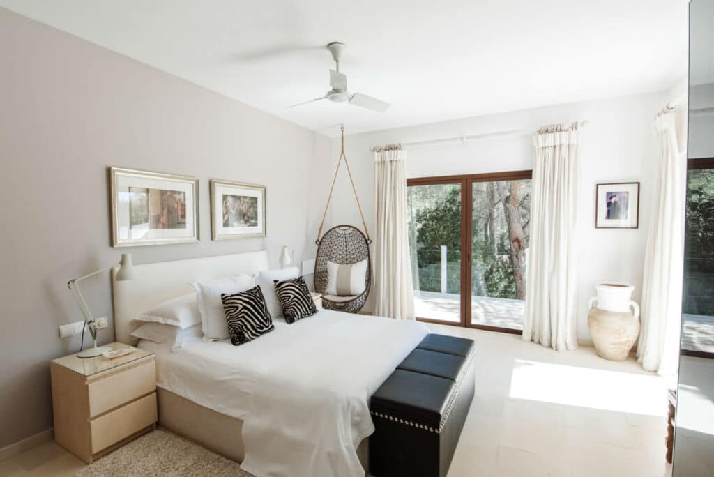 Bright bedroom with a white bed, zebra-patterned pillows, and beige walls evokes a luxury travel vibe. A wicker hanging chair adds charm. Large windows with sheer curtains offer views of greenery. A black bench sits at the foot of the bed, and framed pictures adorn the walls. David Pearson Travel