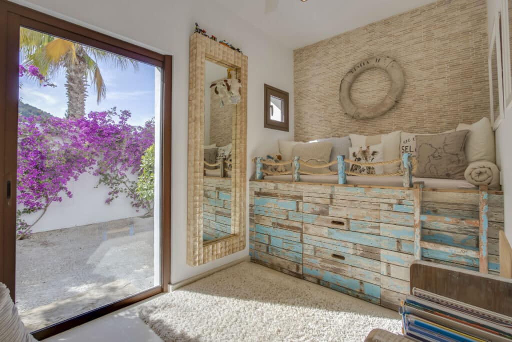 Cozy room with rustic, distressed wooden furniture and a cushioned seating area. A large mirror leans against the wall. Bright bougainvillea is visible through a glass door, adding a splash of color. For those seeking luxury travel vibes, a lifebuoy decor hangs elegantly on the wall. David Pearson Travel