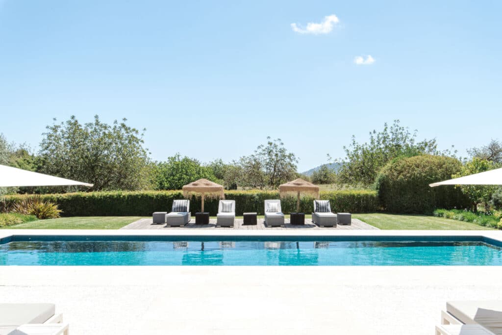 A serene outdoor pool with clear blue water is surrounded by white lounge chairs under straw umbrellas, offering the perfect luxury travel escape. Green hedges and trees border the area with a clear blue sky above, creating an idyllic retreat. David Pearson Travel