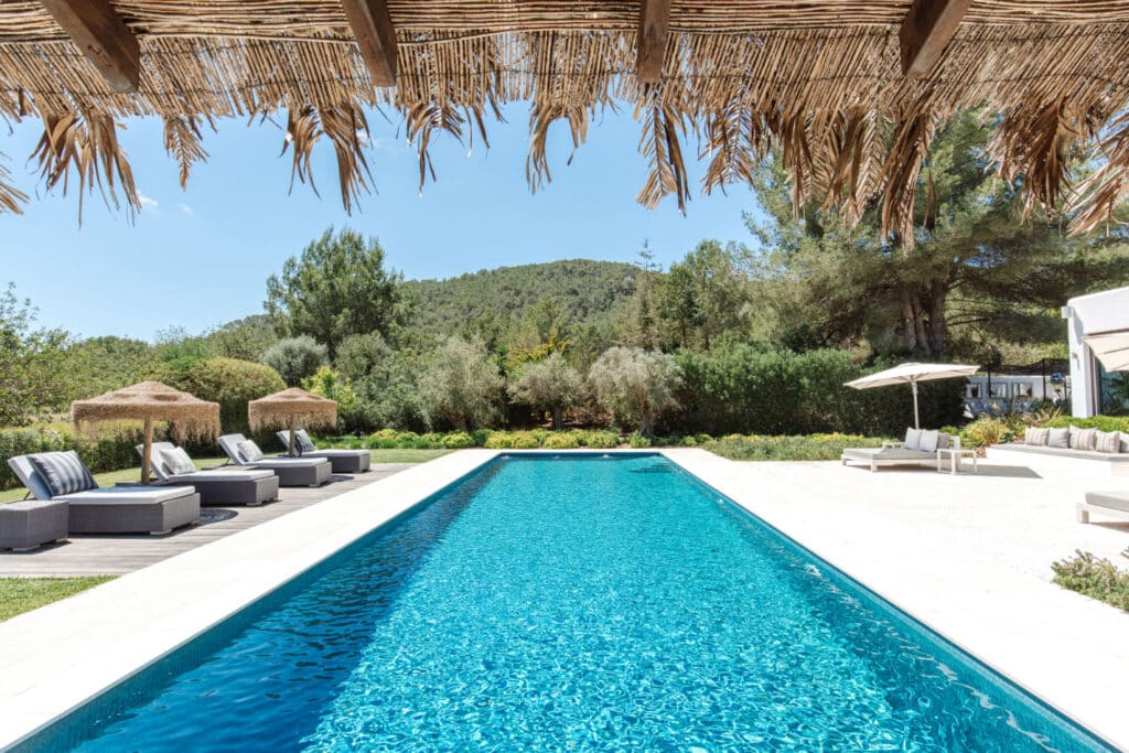 A long, rectangular swimming pool epitomizing luxury travel is surrounded by lounge chairs and umbrellas on a sunny day. Thatched roof fringes hang in the foreground, while lush greenery and a hill paint the background under a clear blue sky. David Pearson Travel
