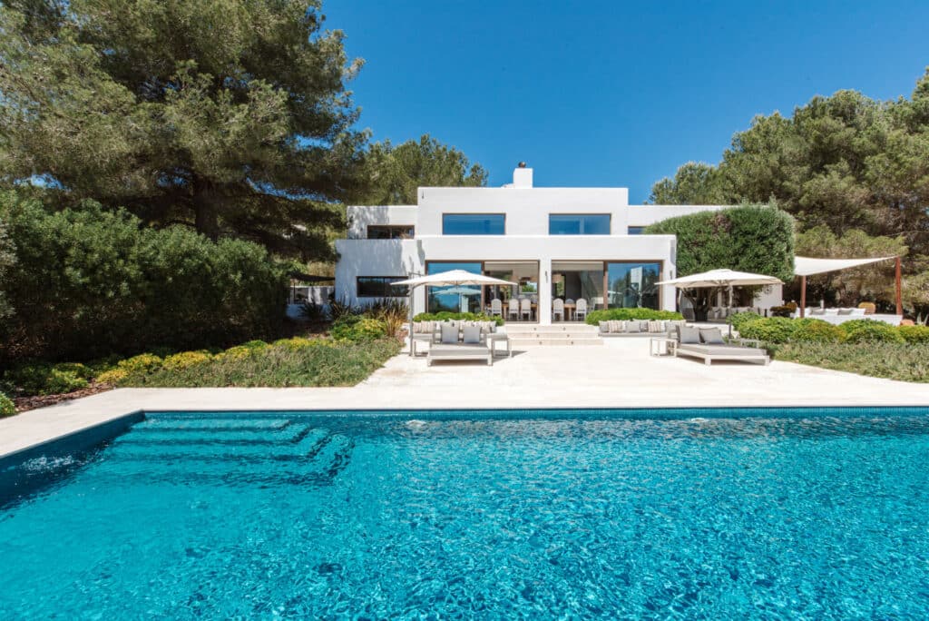 Discover a luxury travel haven in this modern villa with a pristine white facade and expansive windows. Nestled among trees, it overlooks a rectangular swimming pool with clear blue water. The serene setting is completed by a bright, cloudless sky and inviting outdoor seating. David Pearson Travel