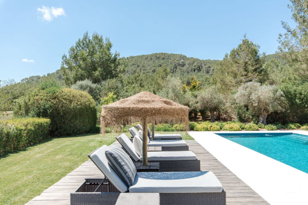 Sun loungers with straw parasols adorn a wooden deck beside a rectangular pool, epitomizing luxury travel. The serene backdrop of lush greenery and distant hills under a clear blue sky enhances this tranquil retreat. David Pearson Travel