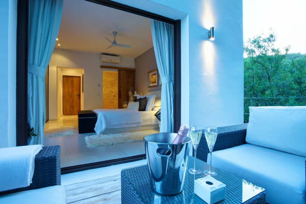 A modern bedroom with open curtains reveals a cozy interior, perfect for luxury travel enthusiasts, featuring a bed with white bedding and a ceiling fan. Outside, on a balcony, theres a wicker sofa and a table with a champagne bucket and two flutes, framed by serene greenery in the background. David Pearson Travel