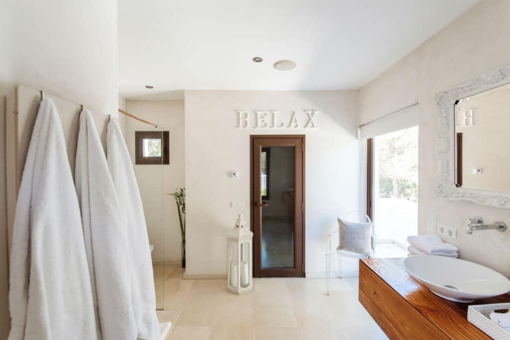 Elegant bathroom with a spa-like ambiance, perfect for luxury travel. Features include white robes on hooks, a large mirror with a decorative frame, and a modern sink on a wooden counter. A door leads outside, where RELAX is tastefully displayed above. David Pearson Travel