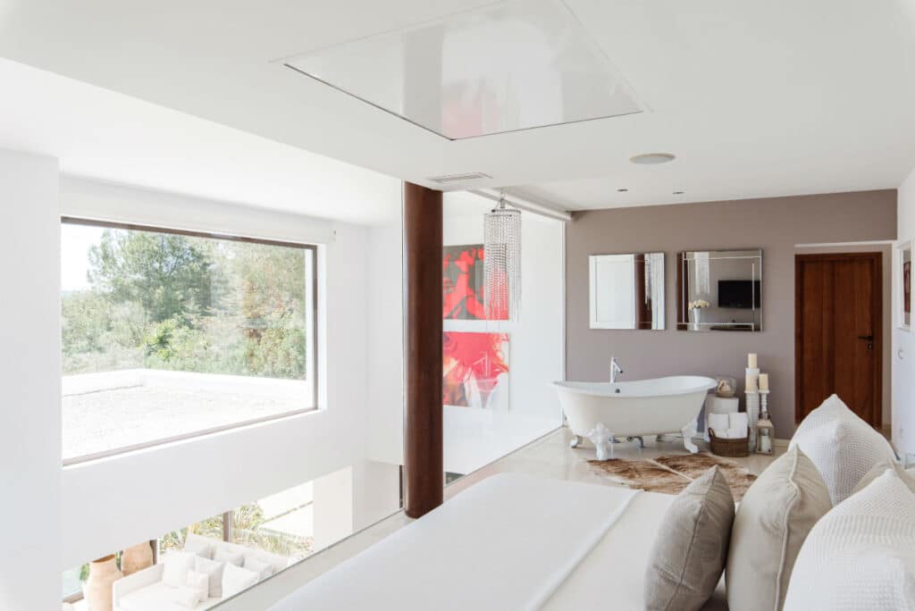 Modern bedroom with large window view of trees, featuring a white freestanding bathtub near a brown accent wall. Perfect for those who appreciate luxury travel, it boasts a bed with white linens in the foreground and a glass-enclosed shower near contemporary wall art. David Pearson Travel