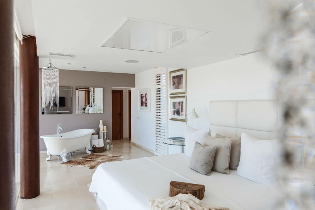 A luxurious bedroom, perfect for luxury travel, features a king-sized bed with white linens and beige pillows. A freestanding bathtub sits in the corner, surrounded by decorative candles. The room boasts neutral tones, framed art on the walls, and large windows with sheer curtains. David Pearson Travel