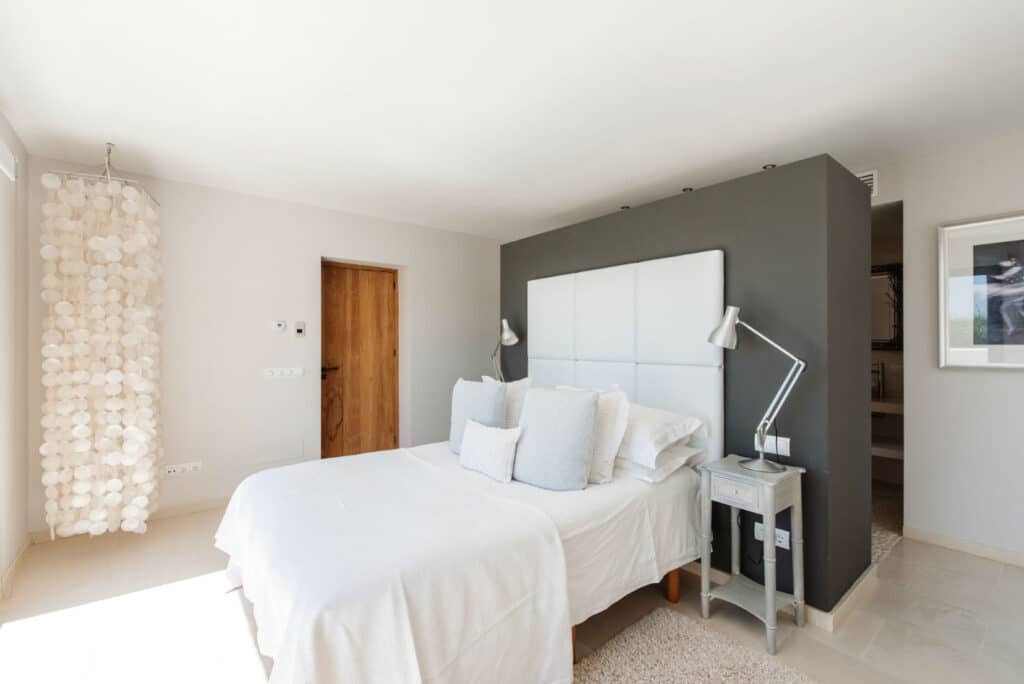A modern bedroom exuding luxury travel vibes features a white bed against a gray accent wall, adorned with plush white pillows. Flanked by two sleek lamps on side tables, the room is bathed in natural light from a window, with an elegant decorative hanging feature on the left. David Pearson Travel