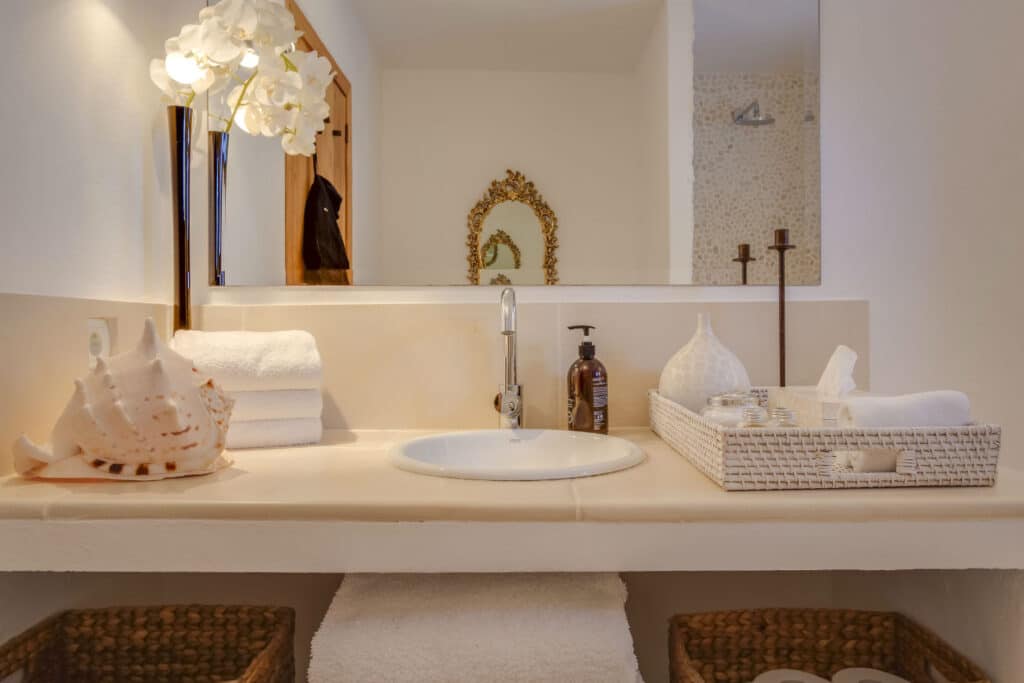 Elegant bathroom sink area with a white countertop, featuring a round sink, white towels, a decorative conch shell, and an orchid plant. A tray holds a soap dispenser and decorative items. A mirror with a golden frame hangs on the wall behind, evoking the sophistication of luxury travel. David Pearson Travel