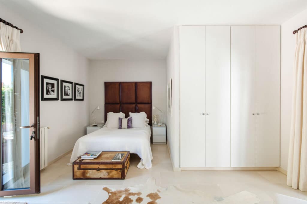 Bright bedroom with a cozy double bed featuring a brown padded headboard, perfect for those seeking a touch of luxury travel at home. White walls and wardrobes enhance the airy feel, while a wooden trunk serves as a coffee table on a beige rug. Three framed photographs decorate the wall beside the bed. David Pearson Travel