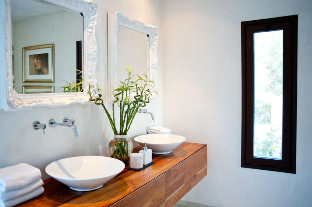 Modern bathroom exuding luxury travel vibes, with two white vessel sinks on a wooden vanity. Ornate mirrors hang above each sink, while a potted plant with tall green stalks adds charm. A narrow window and framed artwork enhance the opulent ambiance. David Pearson Travel