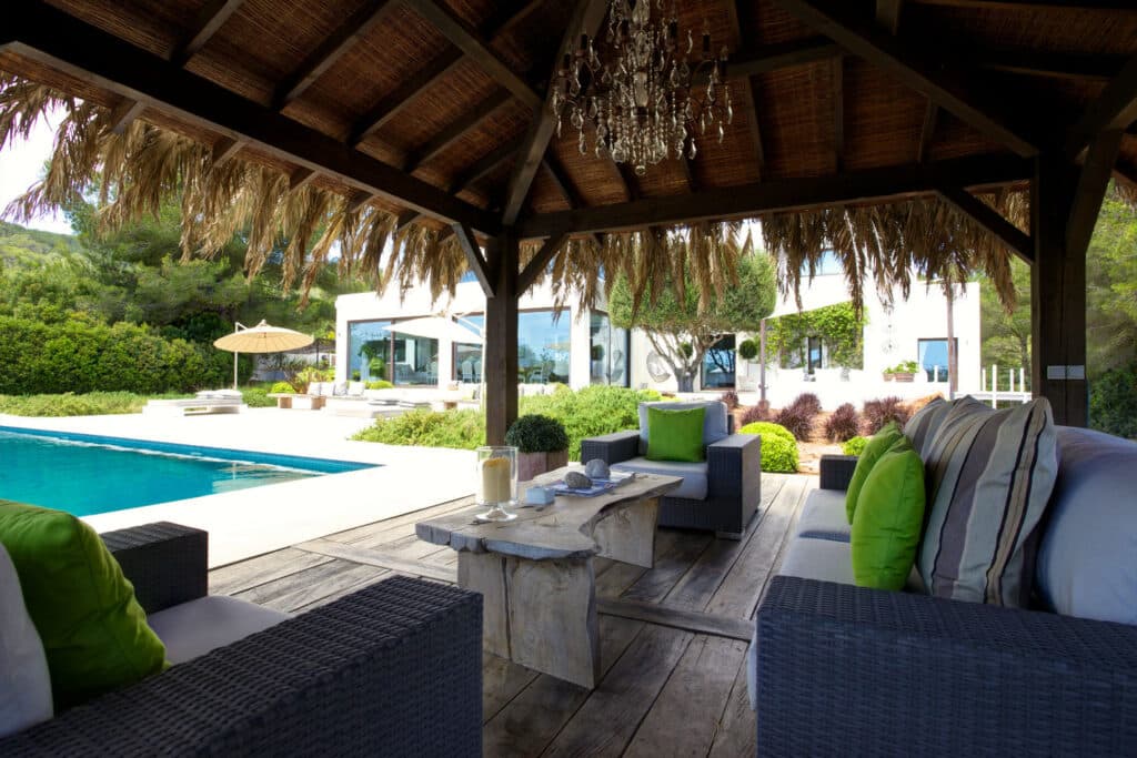 A luxurious outdoor patio perfect for luxury travel experiences features wicker sofas with green and white cushions under a thatched roof pergola. A stone table holds a candle and decorative items. In the background, a modern villa, lush greenery, and a pool with an umbrella complete the scene. David Pearson Travel