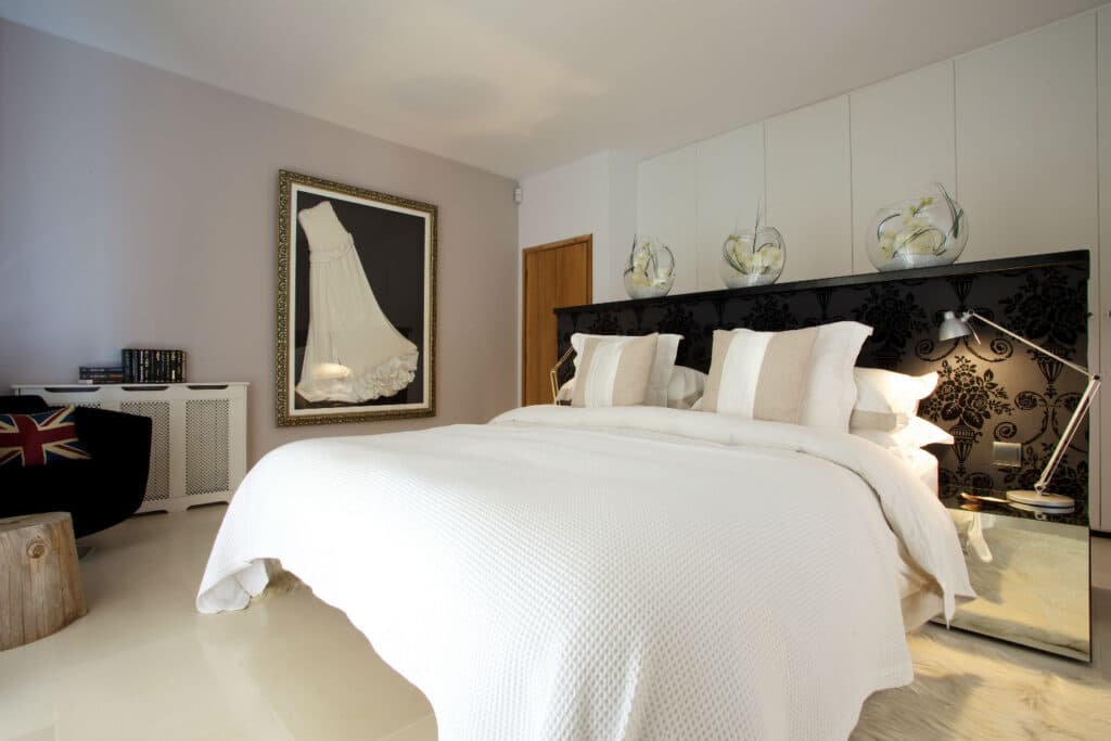 A modern bedroom exuding luxury travel vibes, with a large bed dressed in crisp white linens and plush pillows. Above the headboard, decorative floral vases add charm, while a framed image of a wedding dress graces the wall. A Union Jack cushion rests on a black chair beside a small wooden stool. David Pearson Travel
