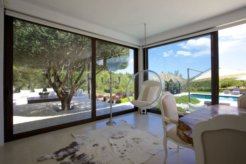 A modern living room epitomizes luxury travel, boasting large glass doors that overlook a lush garden with a tree and pool. The space features a hanging chair, wooden dining table, cowhide rug, and outdoor furniture under a clear blue sky—a perfect retreat for the discerning traveler. David Pearson Travel