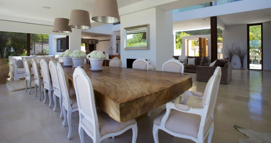 A spacious, modern dining room exudes the elegance of luxury travel with its long wooden table and white upholstered chairs. Overhead, three pendant lights cast a warm glow. The bright room, with large windows showcasing lush greenery, also features a cozy sitting area in the background. David Pearson Travel